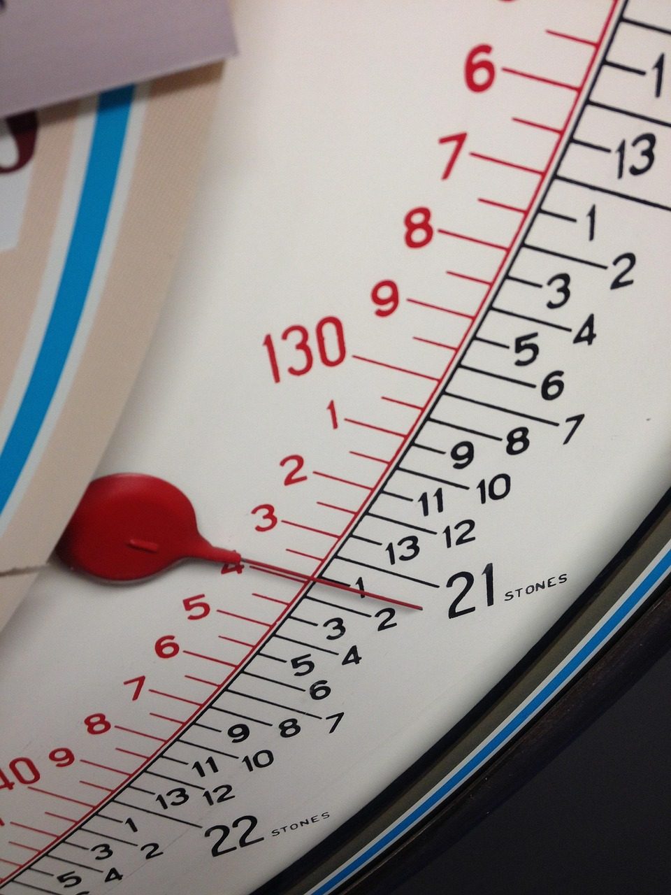 Close-up of a mechanical bathroom scale showing a weight reading around 130 kilograms and 21 stones, with a red pointer indicating the measurement.